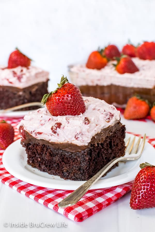 Two white plates with strawberry fudge poke cake slices on them and fresh strawberries on top