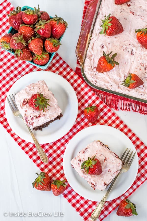 An overhead picture of two slices of strawberry fudge poke cake on white plates on a red and white checkered towel