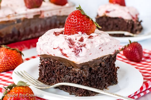 A close up pictures of a slice of strawberry fudge poke cake on a white plate with a fork beside it