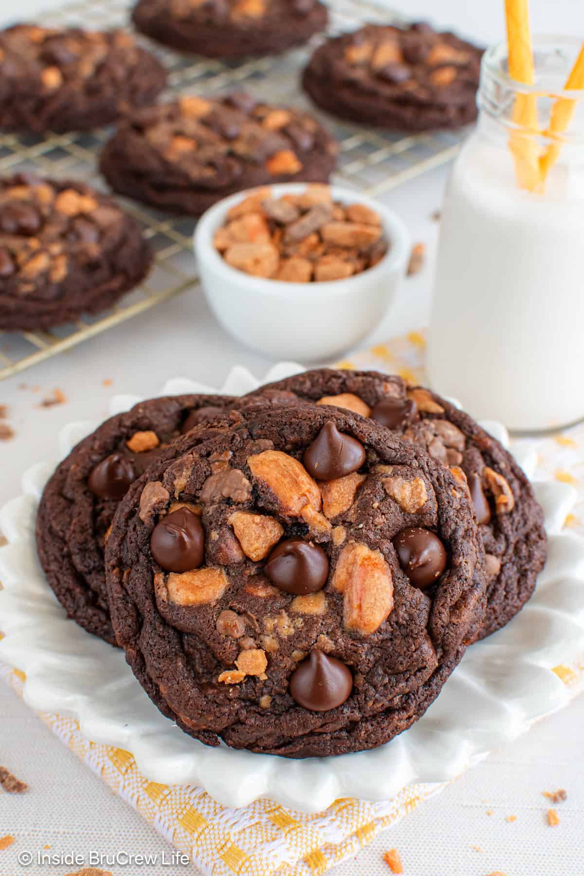 Three chocolate Butterfinger cookies on a white plate with more behind it on a wire rack.