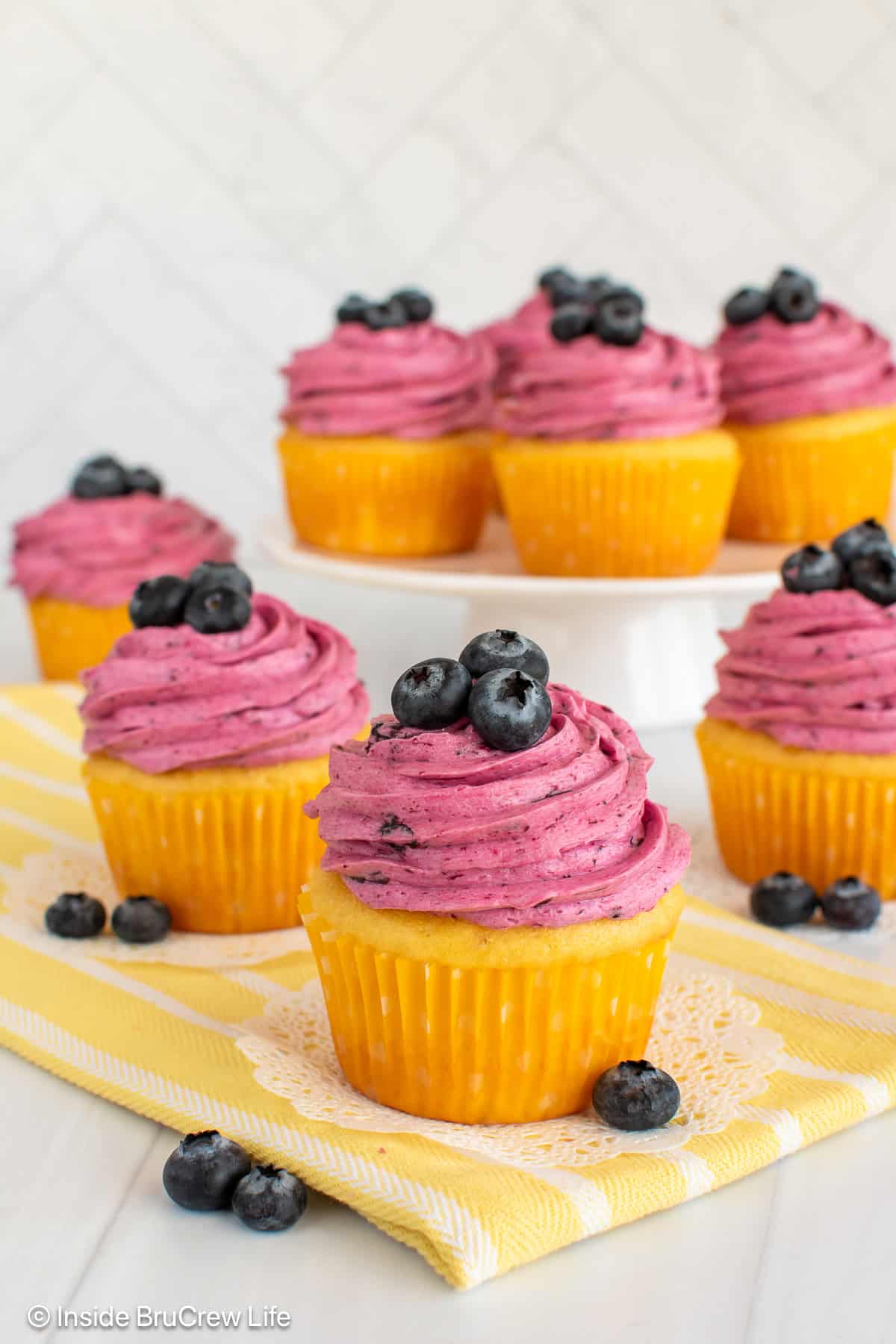 Lemon blueberry cupcakes on a yellow towel and white cake stand.