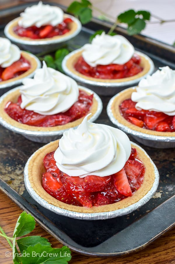 A dark metal baking sheet with six no bake strawberry pies topped with whipped cream on it