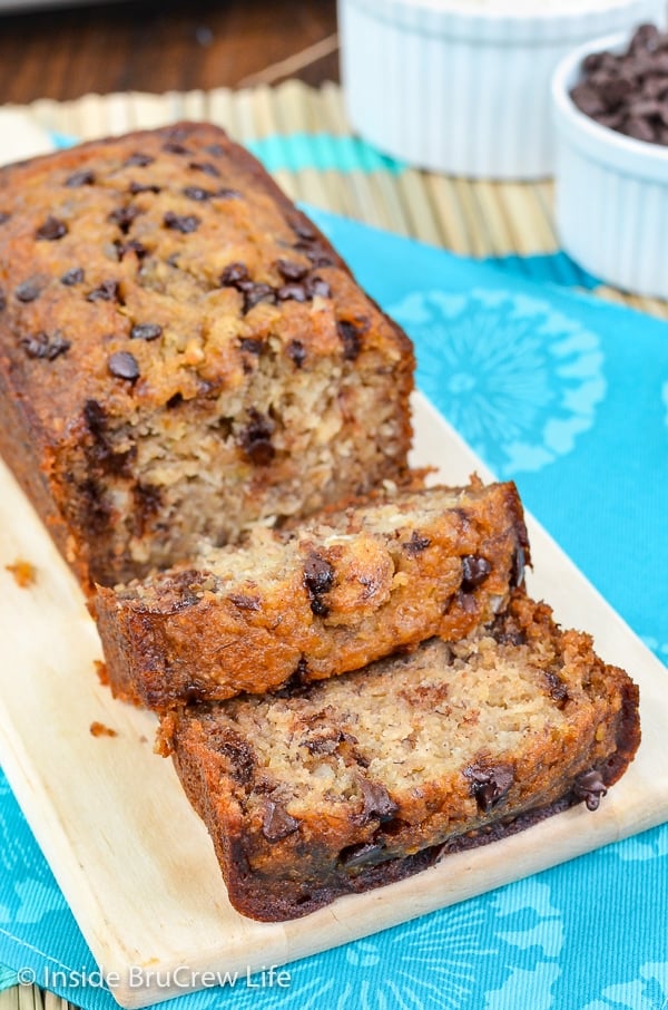 A cutting board with a loaf of healthy chocolate chip banana bread with a few slices falling down in front of it.