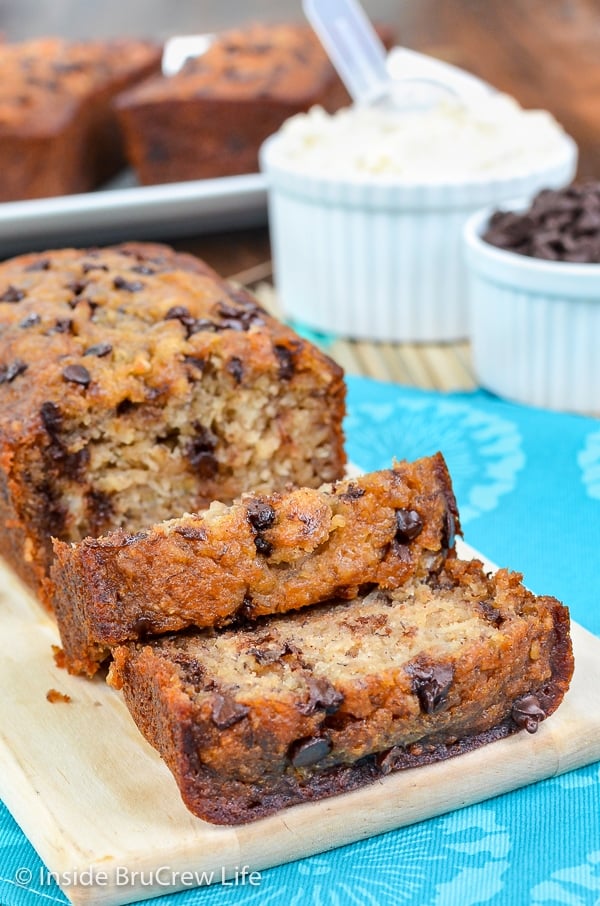 A cutting board with a loaf of healthy chocolate chip banana bread with a few slices falling down in front of it.