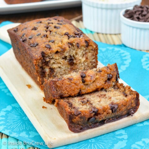 A cutting board with a loaf of healthy chocolate chip banana bread with a few slices falling down in front of it.