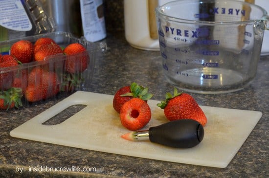 A white cutting board with fresh strawberries and a strawberry corer on it