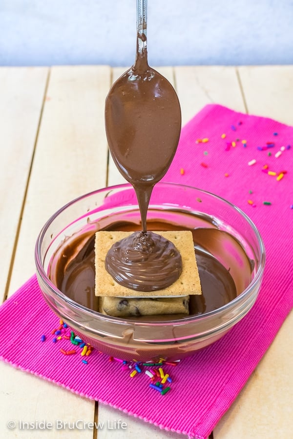 A bowl of melted chocolate on a pink towel with a spoon drizzling chocolate on top of a no bake cookie dough s'mores