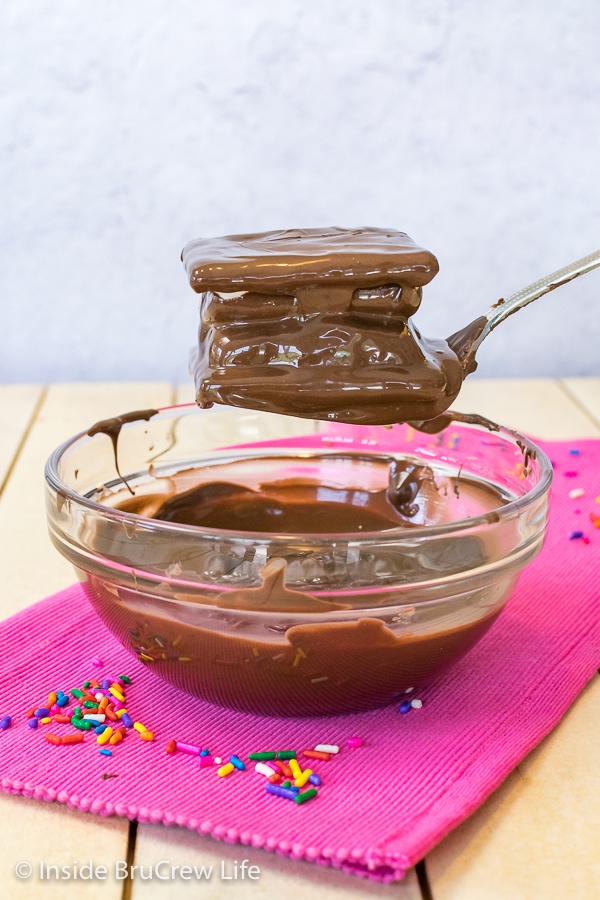 A bowl of melted chocolate on a pink towel with a no bake cookie dough s'mores being lifted out of it with a fork