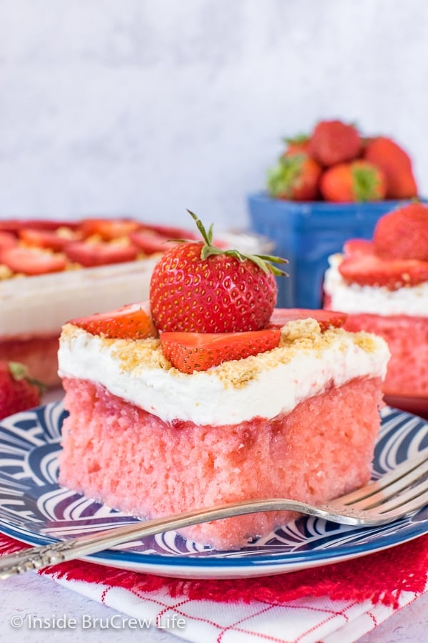 Strawberry Cheesecake Poke Cake - this easy poke cake has strawberry in it and a fluffy cheesecake mousse on top. Make this recipe for summer parties! A blue plate with a square of strawberry cheesecake poke cake with a strawberry on top.