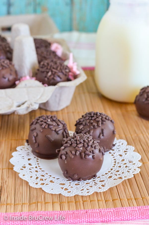 Three chocolate covered butterfinger cookie dough bites topped with chocolate sprinkles on a white doily