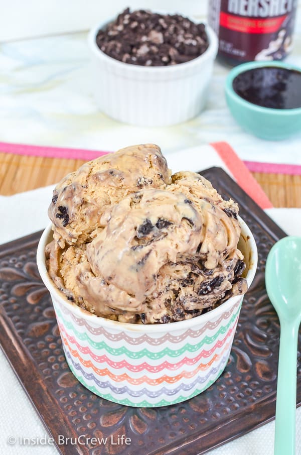 A white paper cup on a brown tile filled with oreo fudge cappucino ice cream and bowls of oreo and hot fudge behind it