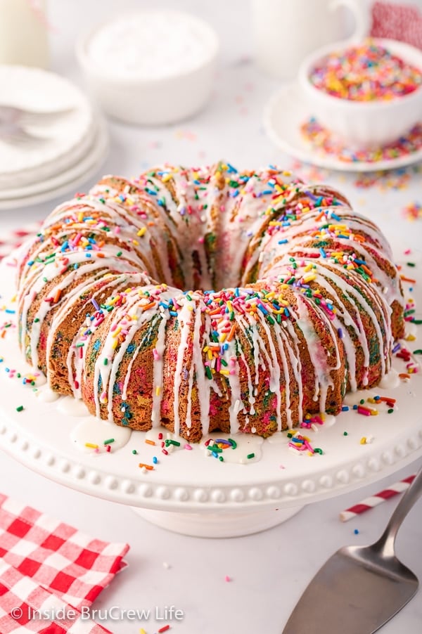 A white cake plate with a bundt cake topped with white glaze and sprinkles on it.