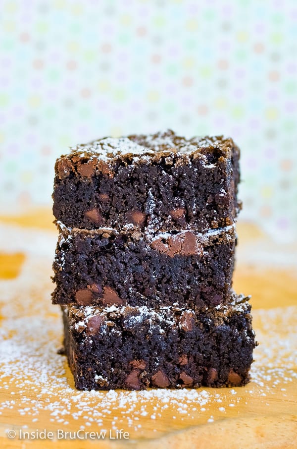 A cutting board with three homemade chocolate chip brownies stacked up and sprinkled with powdered sugar.