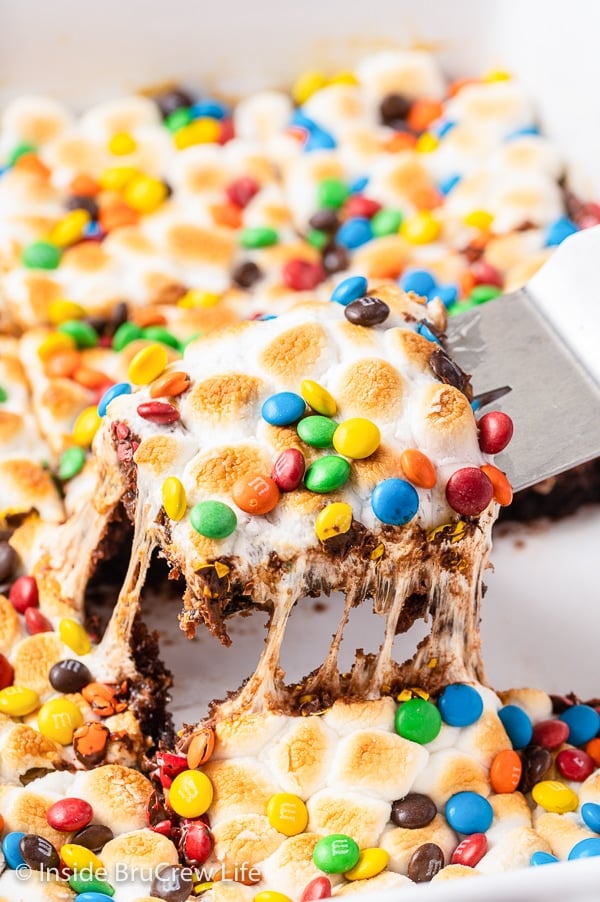 A pan of caramel marshmallow brownies with M&M's and a spatula lifting out a gooey square.