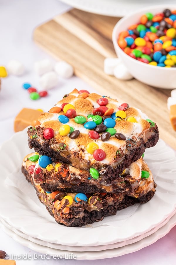 Three marshmallow topped brownies with M&M's stacked on a white plate.