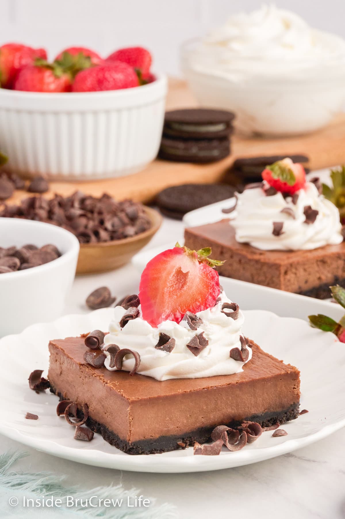 A white plate with a chocolate cheesecake bar topped with whipped cream and strawberries.