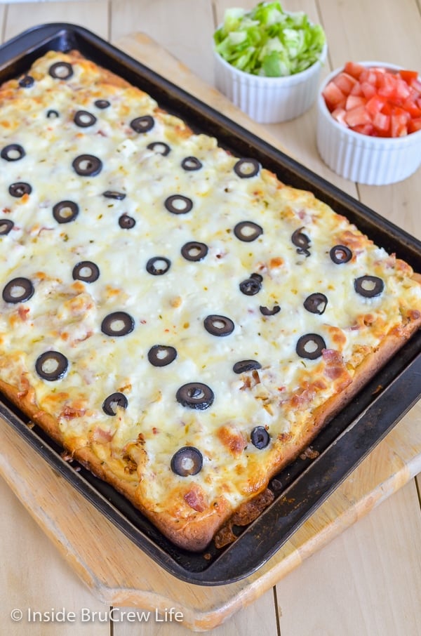 A sheet pan with a cooked ranch chicken bacon club pizza topped with olives on it
