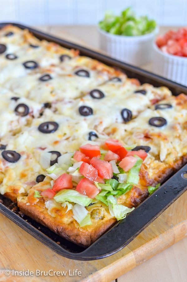 A sheet pan with a cooked chicken bacon club pizza on it and one square topped with lettuce and tomatoes