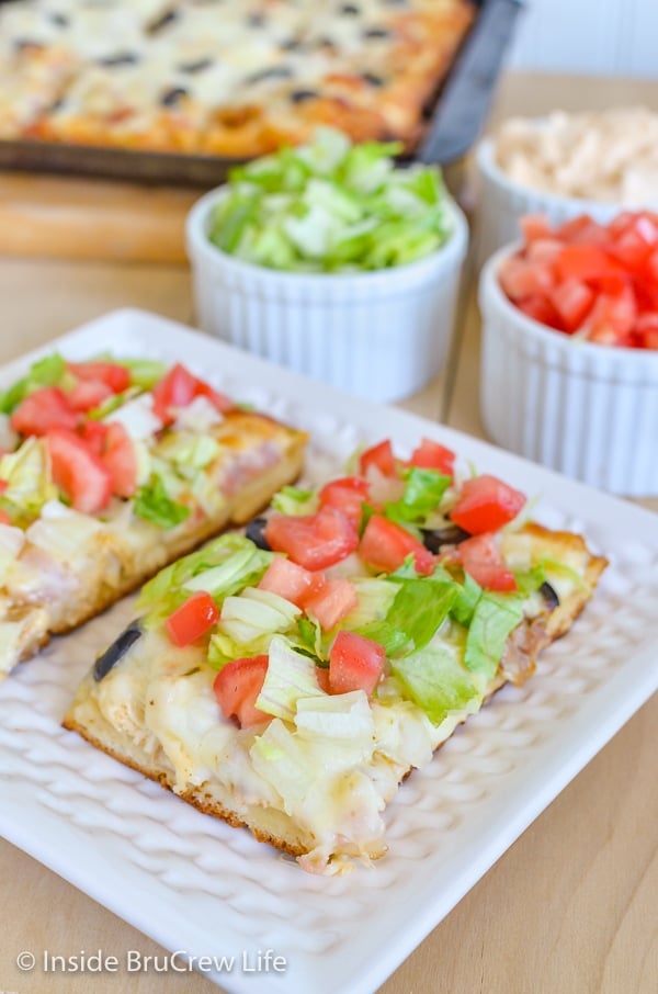 A white plate with two squares of chicken bacon club pizza on it and bowls of veggies behind it