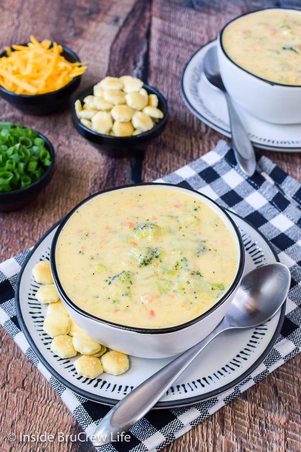 Two white bowls on white plates filled with broccoli cheddar soup and crackers beside the bowls.