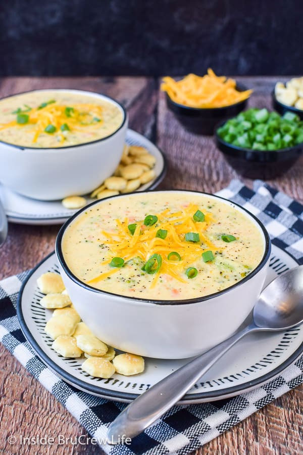 Two white bowls filled with broccoli cheese soup and topped with shredded cheese and green onions.