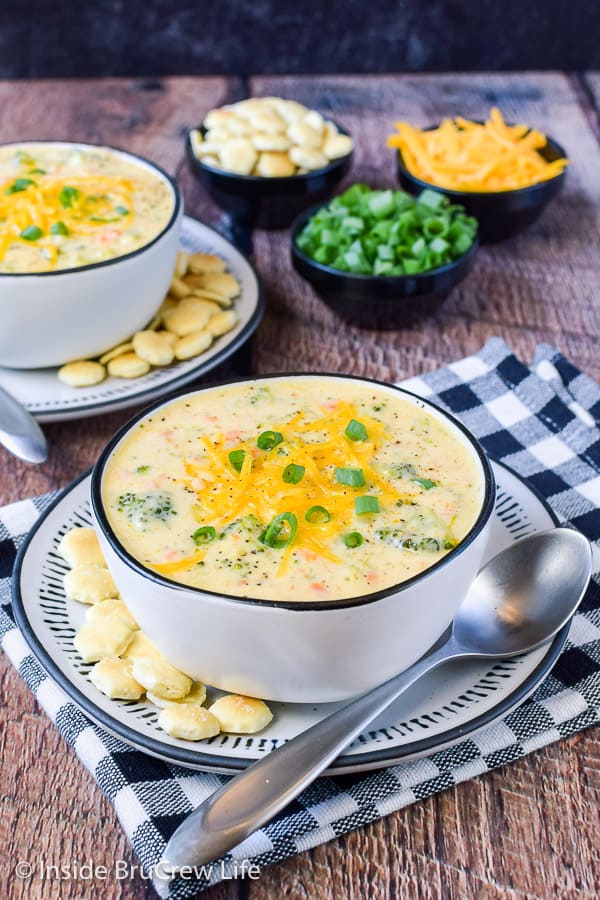 A bowl of broccoli cheese soup topped with cheese and green onions and crackers beside the bowl.