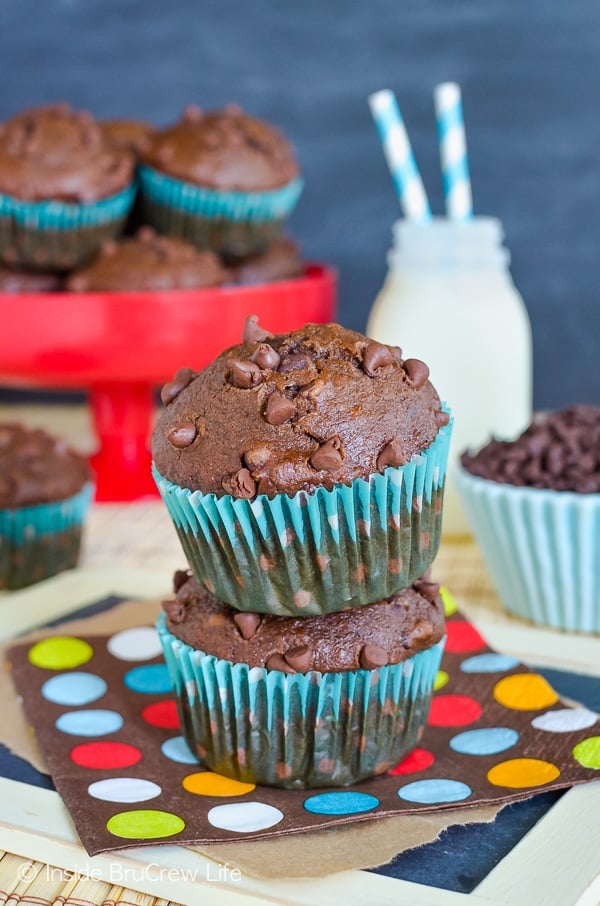 Two double chocolate banana muffins stacked on top of each other on a colorful napkin. More muffins and milk behind them.