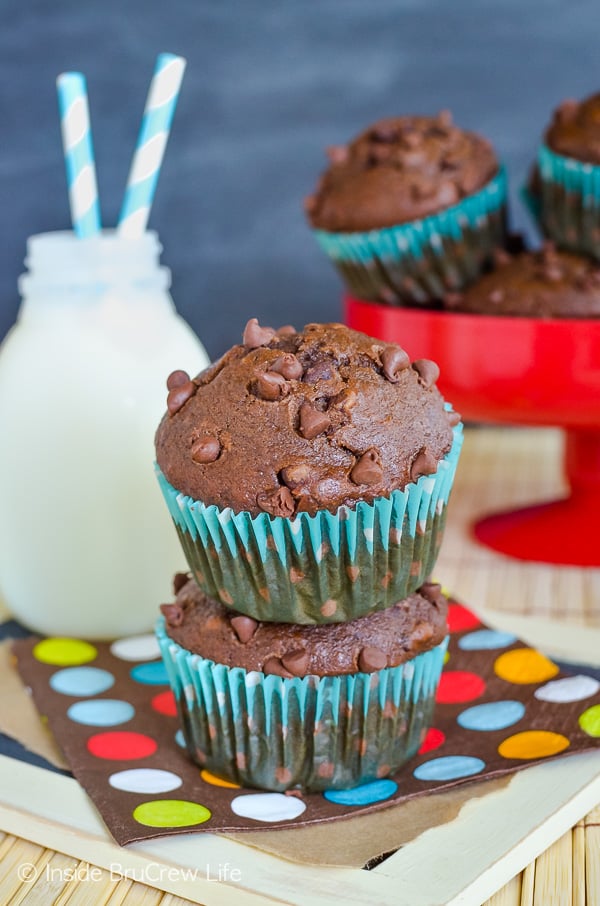 Two chocolate banana muffins with chocolate chips on a colorful napkin. A red plate with more muffins is behind them.