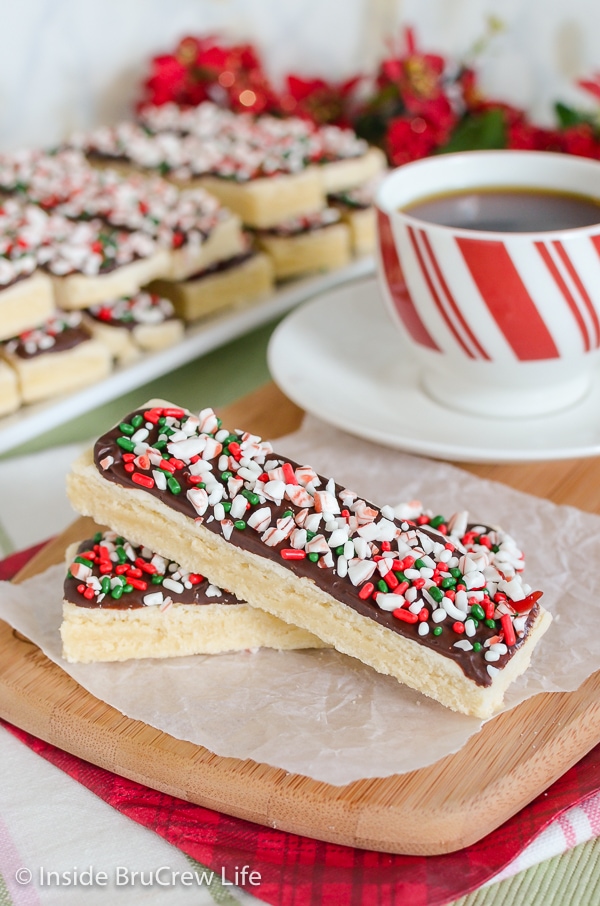 Two shortbread sticks topped with chocolate and candy cane bits on a wooden cutting board.