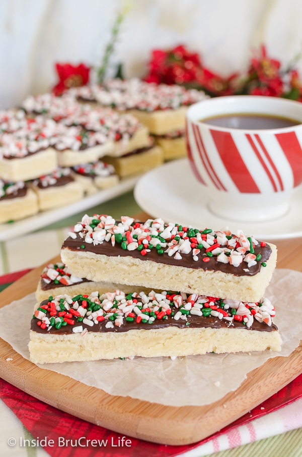 Three shortbread cookies sticks decorated with chocolate and peppermint bits stacked on a wooden cutting board.