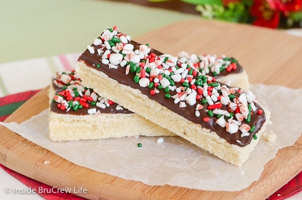 A wooden cutting board with two peppermint shortbread bars decorated with chocolate and candy canes on it.