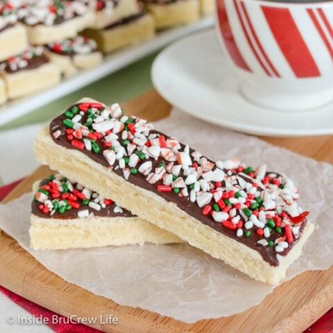 Two shortbread bars topped with chocolate and candy cane bits stacked on a cutting board.