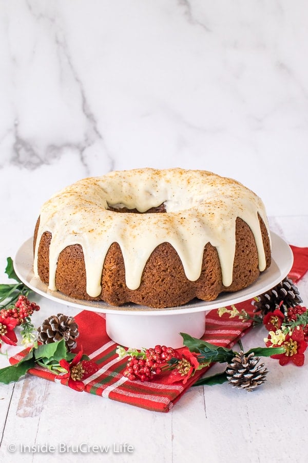 A white cake plate with a full eggnog bundt cake topped with eggnog glaze and nutmeg on it