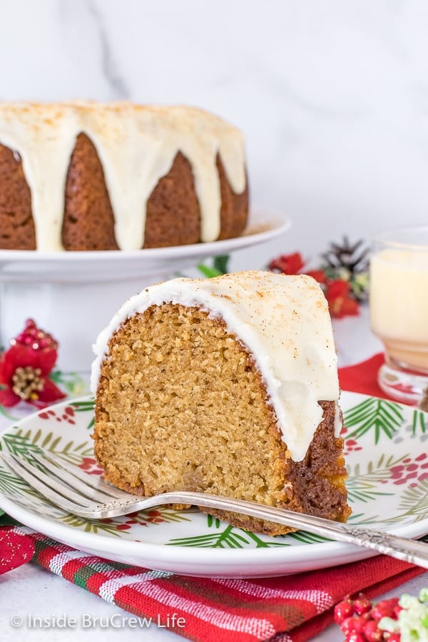 A slice of Eggnog Bundt Cake topped with eggnog glaze on a white plate with more cake on a cake plate behind it