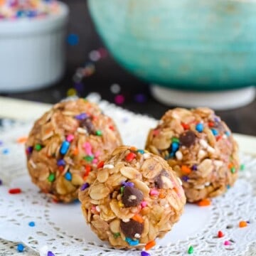 Three peanut butter granola bites on a white doily with a bowl of bites behind them