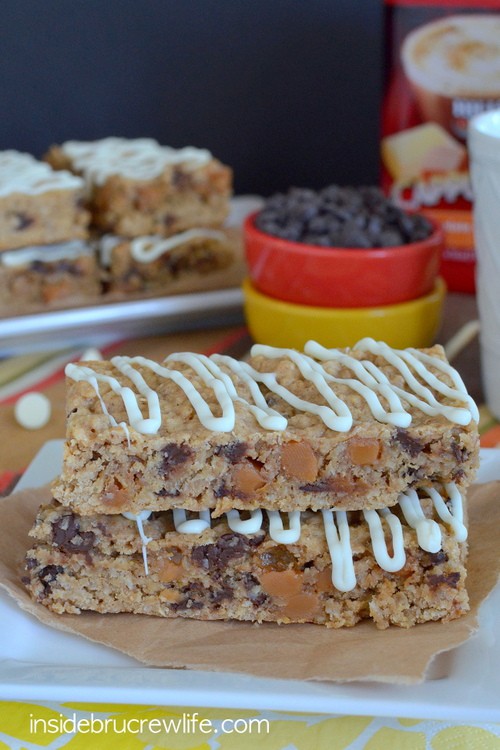 Two caramel granola bars drizzled with white chocolate stacked on a white plate with more behind it