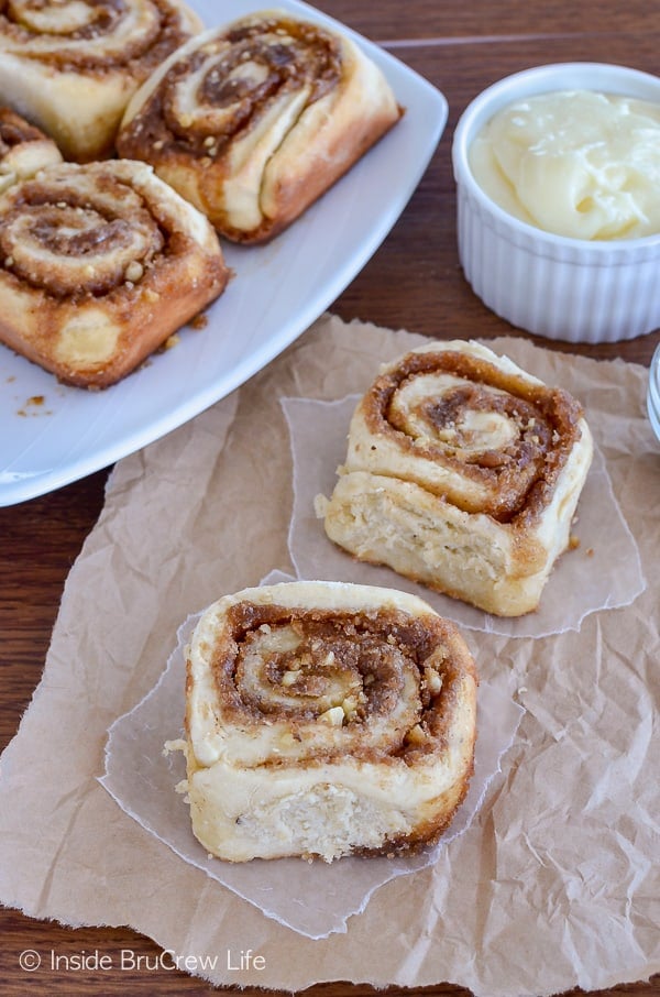 Two baked banana nut cinnamon rolls on parchment with a plate of rolls behind them.