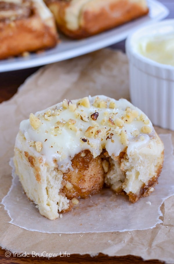 A banana cinnamon roll with frosting and nuts on a piece of parchment with a bite take out of the front.