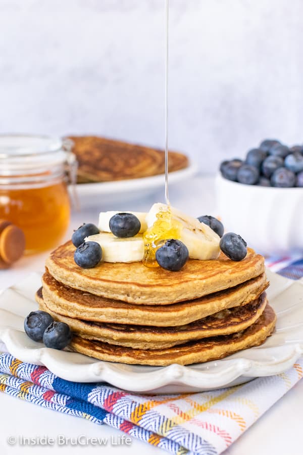 A stack of banana oatmeal pancakes on a white plate with banana slices and blueberries and drizzle of honey coming down on top