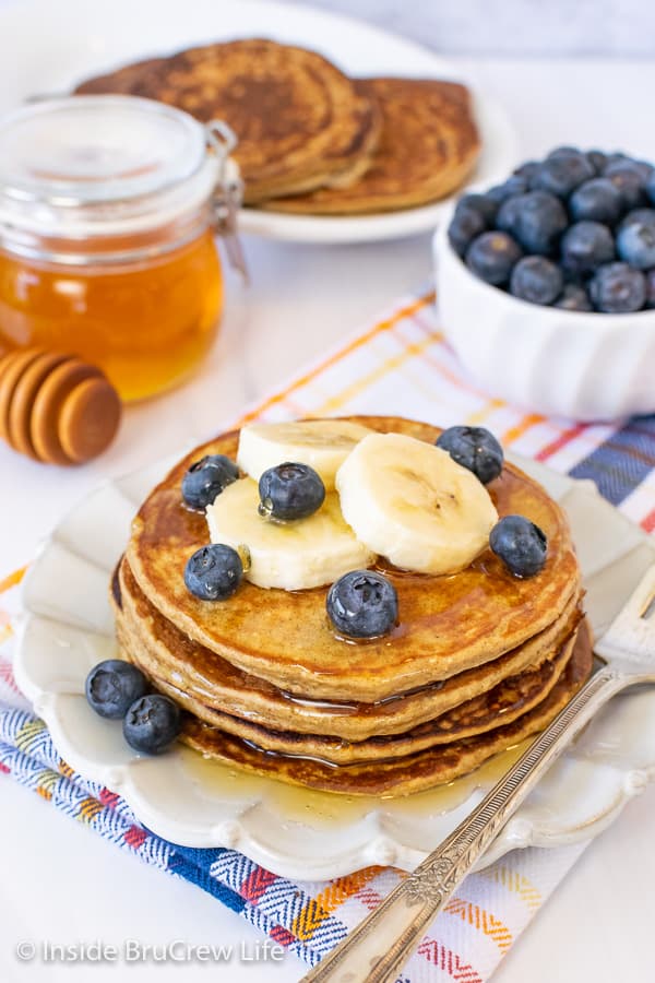 A stack of banana oatmeal pancakes on white plate with bananas and blueberries on top and a bowl of blueberries and a jar of honey behind it