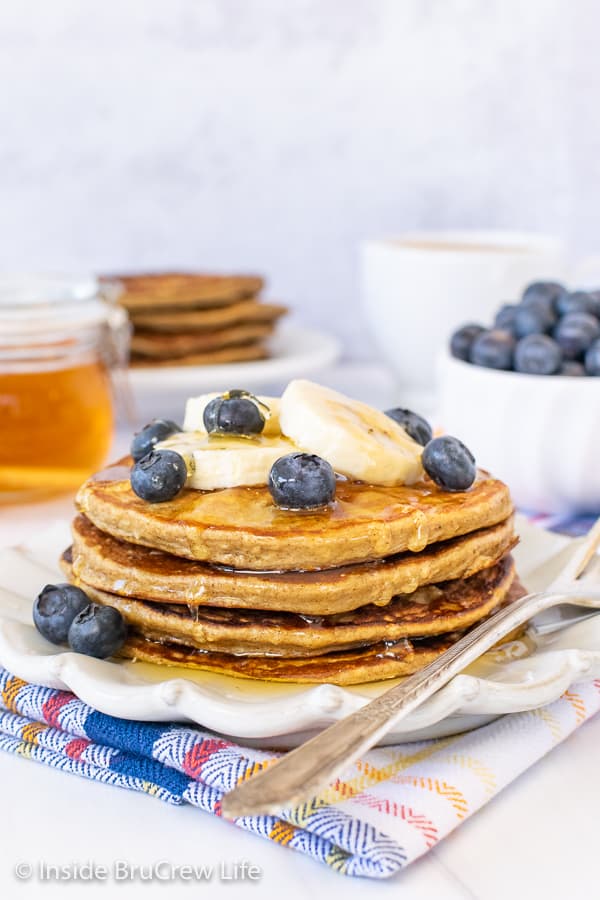 A stack of four banana oatmeal pancakes on a white plate topped with bananas, blueberries, and honey