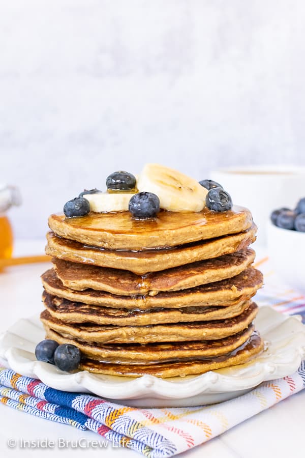 A stack of eight banana oatmeal pancakes on a white plate with banana slices, blueberries, and honey drizzles on top