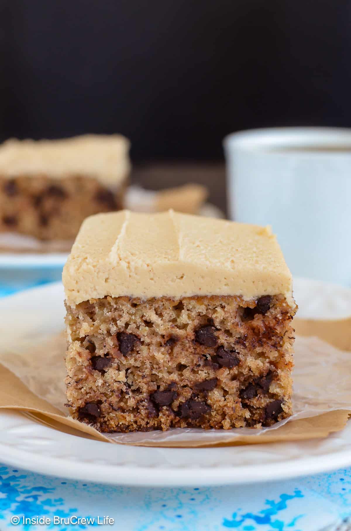Two squares of banana cake with peanut butter frosting on white plates.