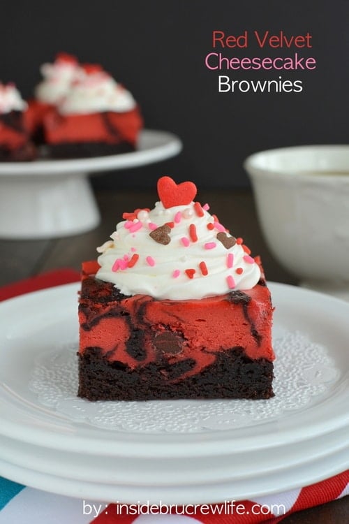 Stack of white plates with Red Velvet Cheesecake Brownies on it with a white cake stand in the background with more brownies