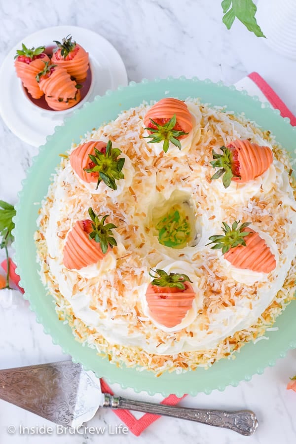 Above picture of a bundt cake topped with frosting, toasted coconut, and orange dipped strawberries on a light green cake plate.