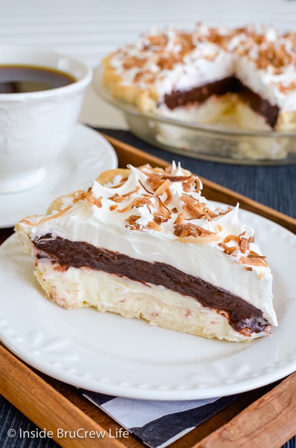 A white plate on a wood tray with a slice of coconut chocolate pie with cool whip and toasted coconut on it and pie plate behind it