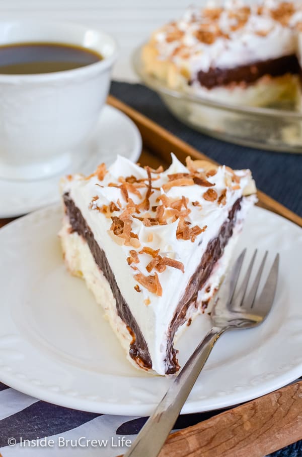 A slice of chocolate coconut pudding pie topped with Cool Whip and toasted coconut on a white plate with a fork beside it