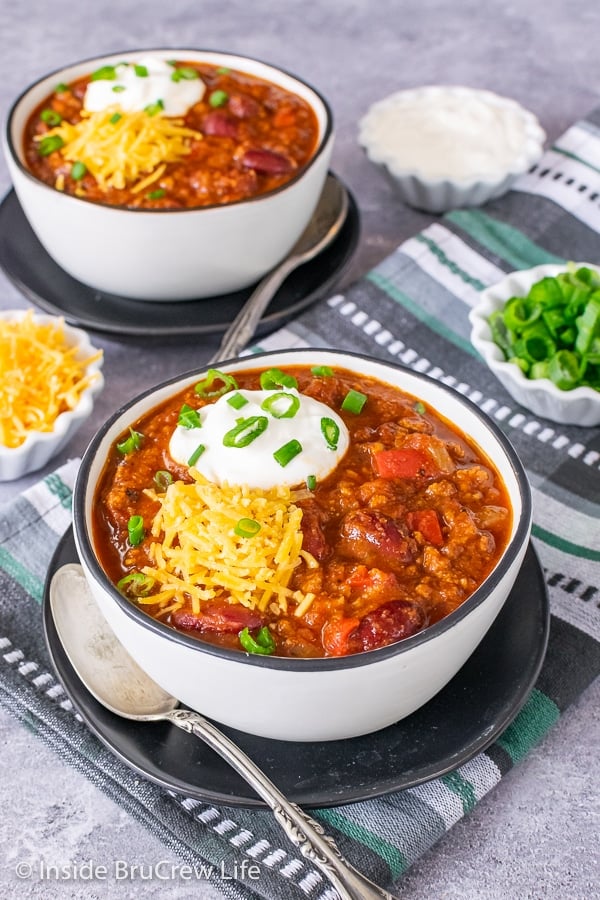 Two white bowls on black plates filled with Dr. Pepper Chili and topped with shredded cheese, sour cream, and green onions