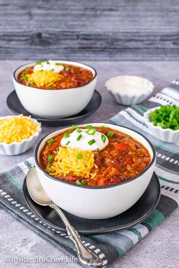 Two white bowls on black plates filled with Sweet and Spicy Dr. Pepper Chili and topped with shredded cheese, sour cream, and green onions