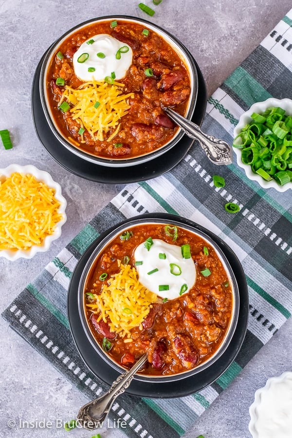 Overhead picture of two bowls on black plates filled with Dr. Pepper Chili and topped with diced green onions, shredded cheddar cheese, and sour cream